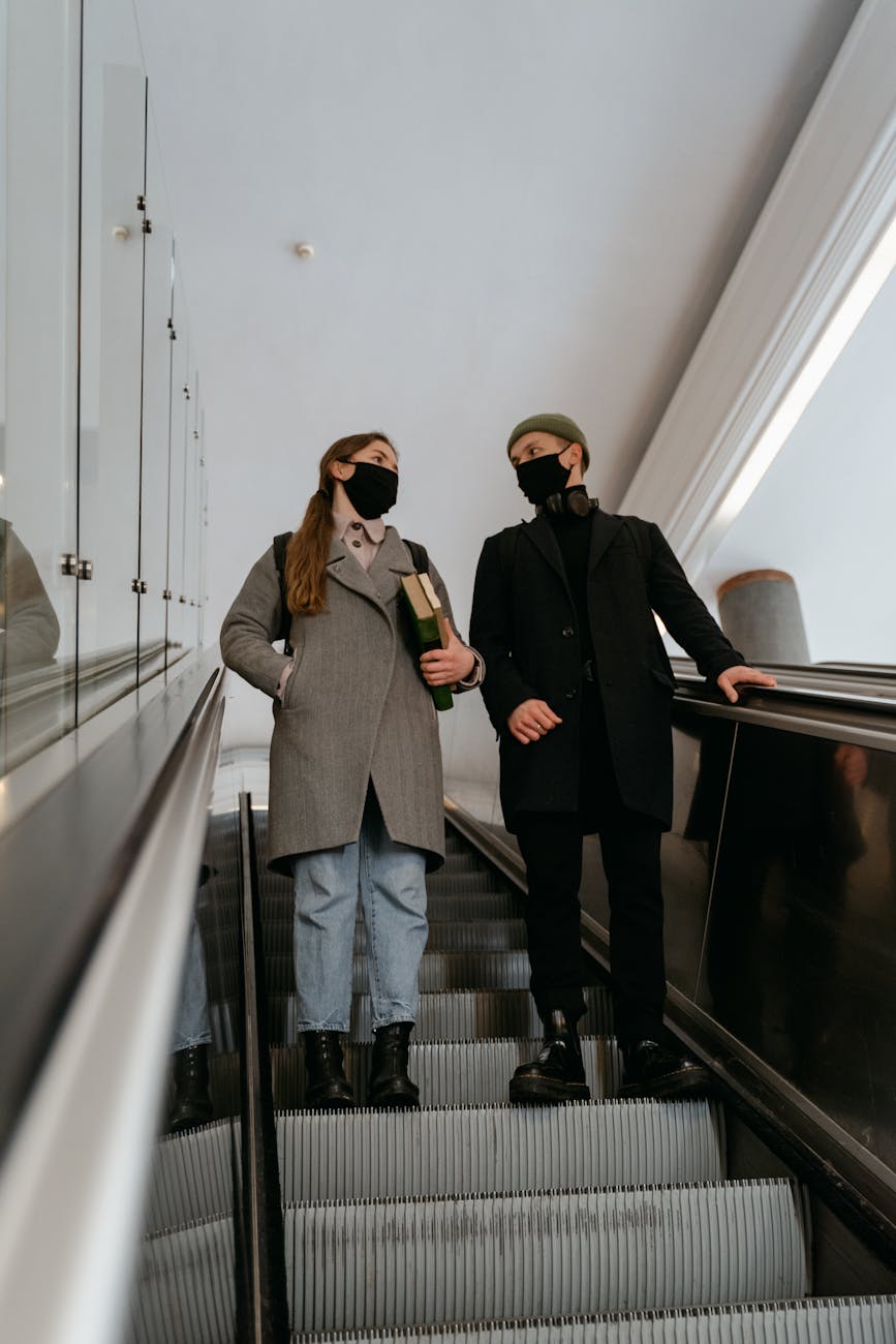 Two students wearing face masks while descending an escalator indoors, reflecting modern education life.